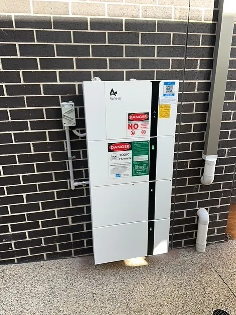 Home battery and inverter installed against a dark brick wall with safety labelling